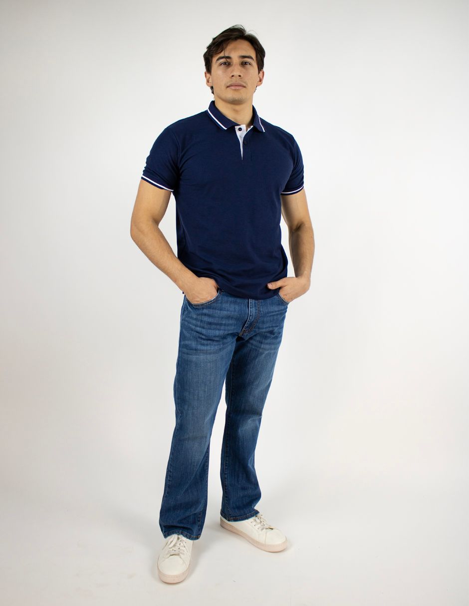 Hombre de pie sobre un fondo blanco, vistiendo un polo azul marino, vaqueros azules y zapatillas blancas.