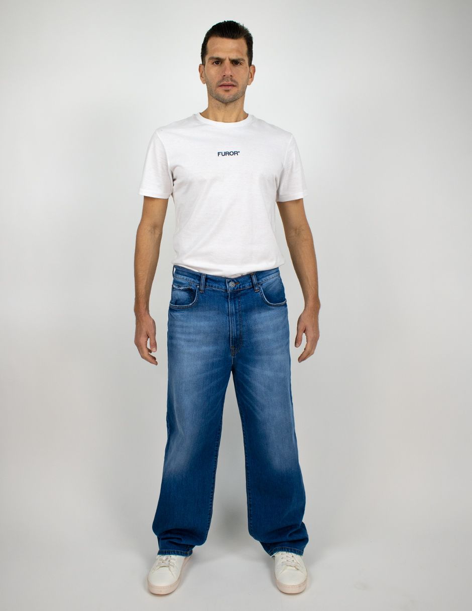 Hombre de pie frente a un fondo liso, vistiendo una camiseta blanca, vaqueros azules y zapatillas blancas.