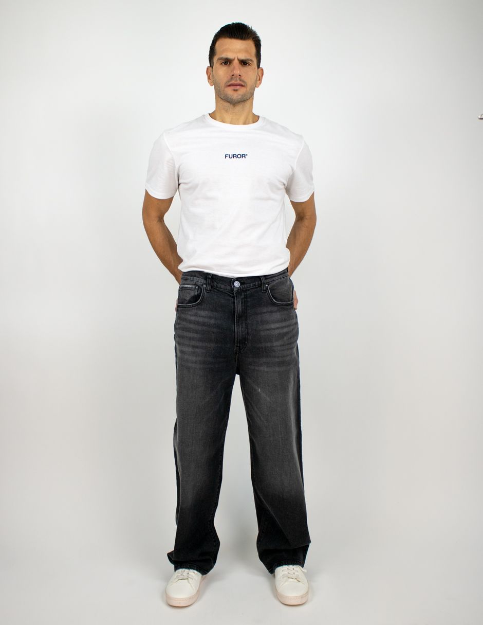 Hombre de pie sobre un fondo blanco, vistiendo una camiseta blanca, vaqueros oscuros y zapatos blancos.
