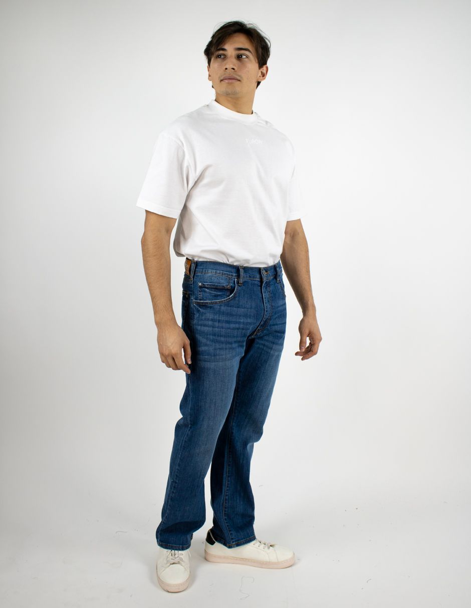 Hombre de pie sobre un fondo blanco, vistiendo una camiseta blanca, vaqueros azules y zapatillas blancas.