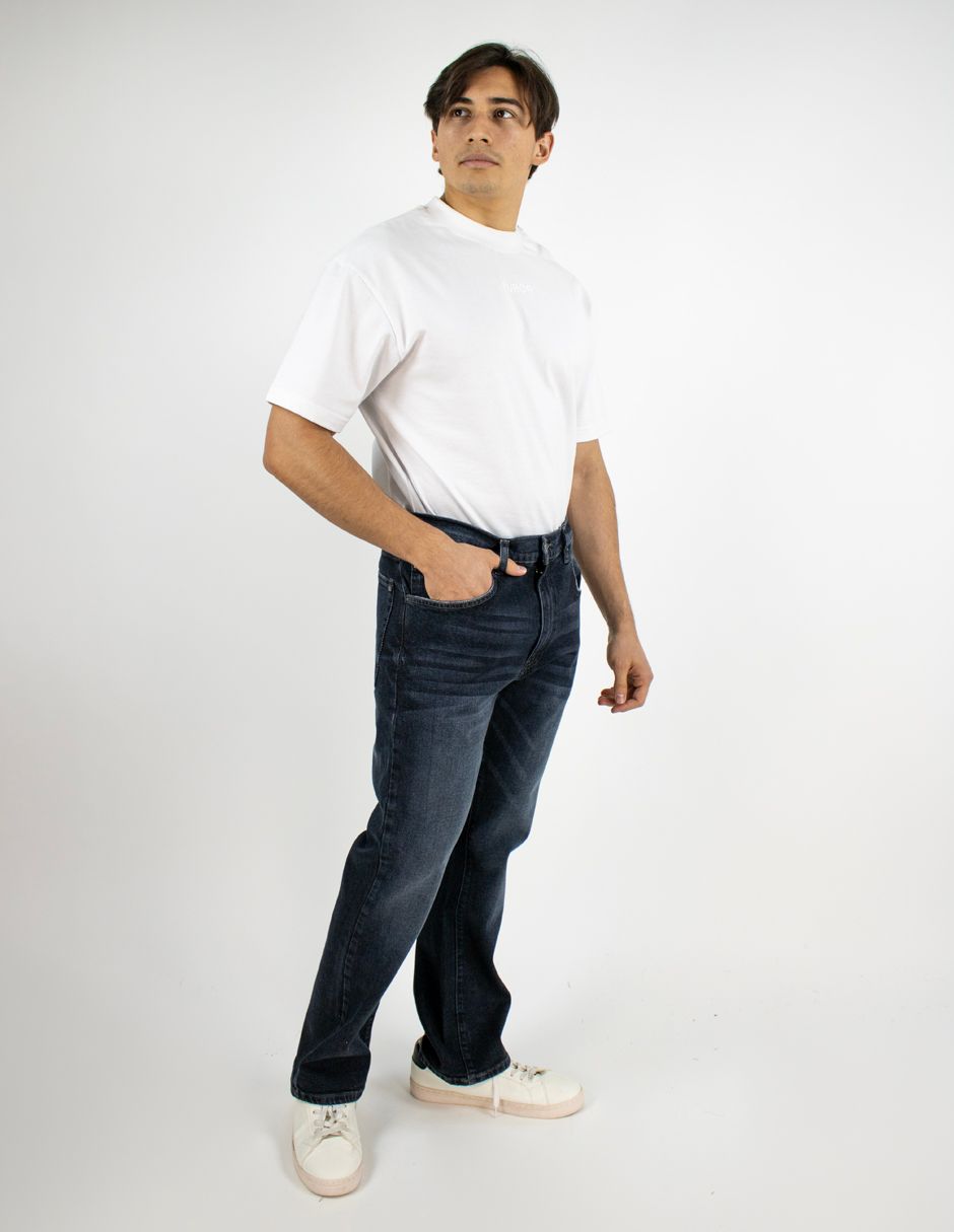 Hombre de pie con camiseta blanca y vaqueros oscuros sobre un fondo blanco liso.