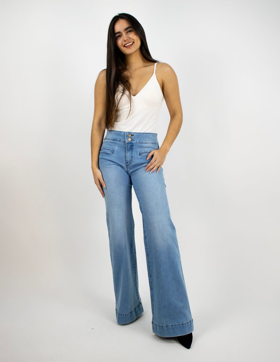 Mujer con top blanco y vaqueros acampanados azul claro posando sobre un fondo blanco liso.