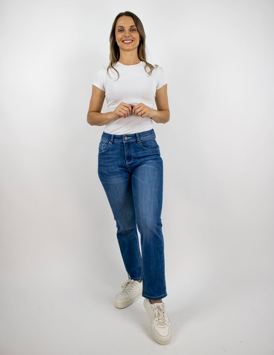 Mujer de pie sobre un fondo blanco, vestida con una camiseta blanca, vaqueros azules y zapatillas blancas.