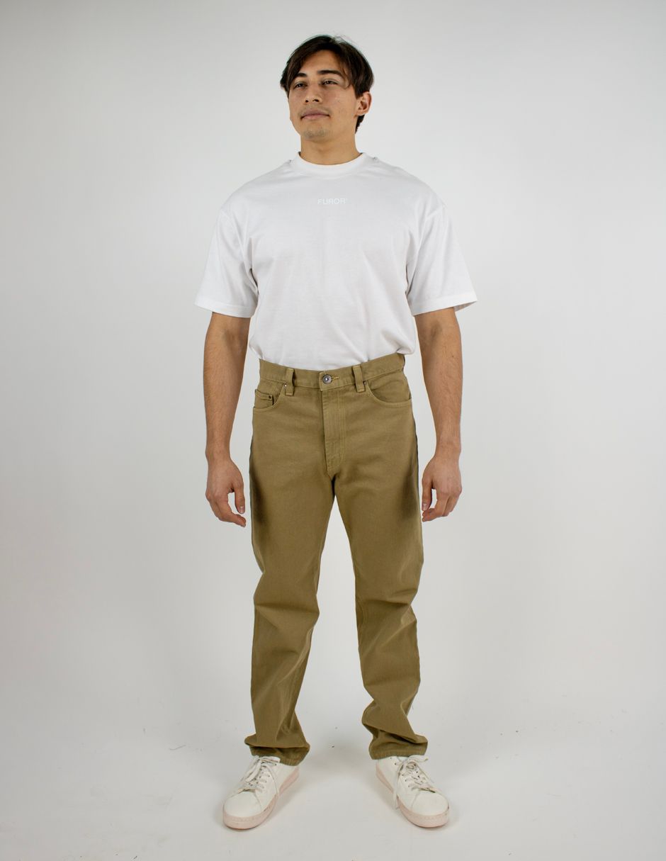 Hombre de pie sobre un fondo blanco liso, vistiendo una camiseta blanca, pantalones verde oliva y zapatillas blancas.
