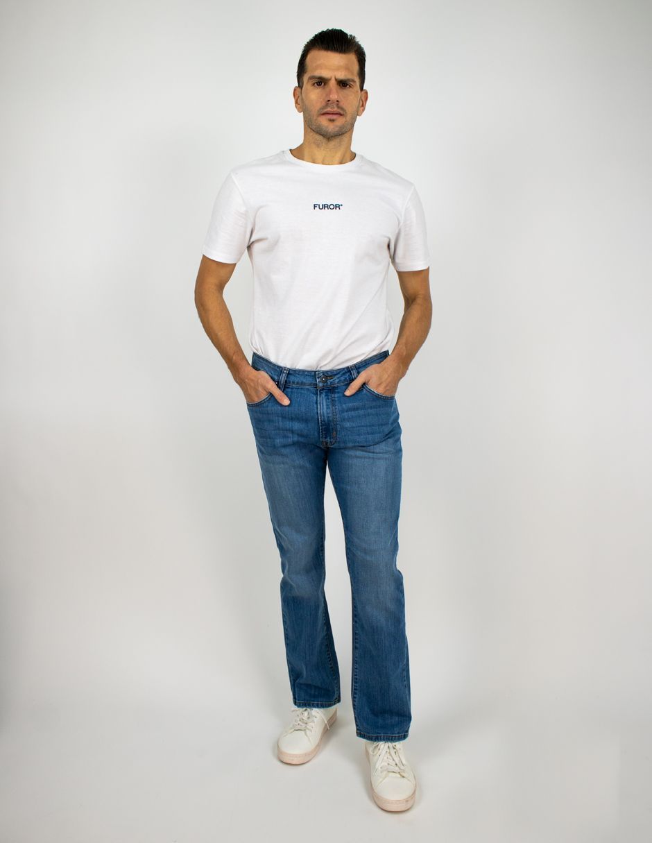 Persona de pie sobre un fondo liso, vestida con una camiseta blanca, vaqueros azules y zapatillas blancas.