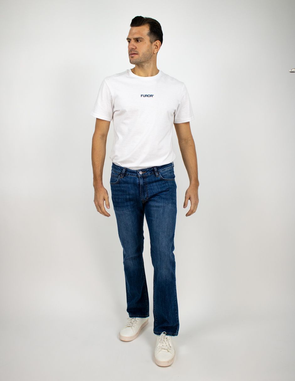 Hombre de pie sobre un fondo blanco, vistiendo una camiseta blanca, vaqueros azules y zapatillas blancas.