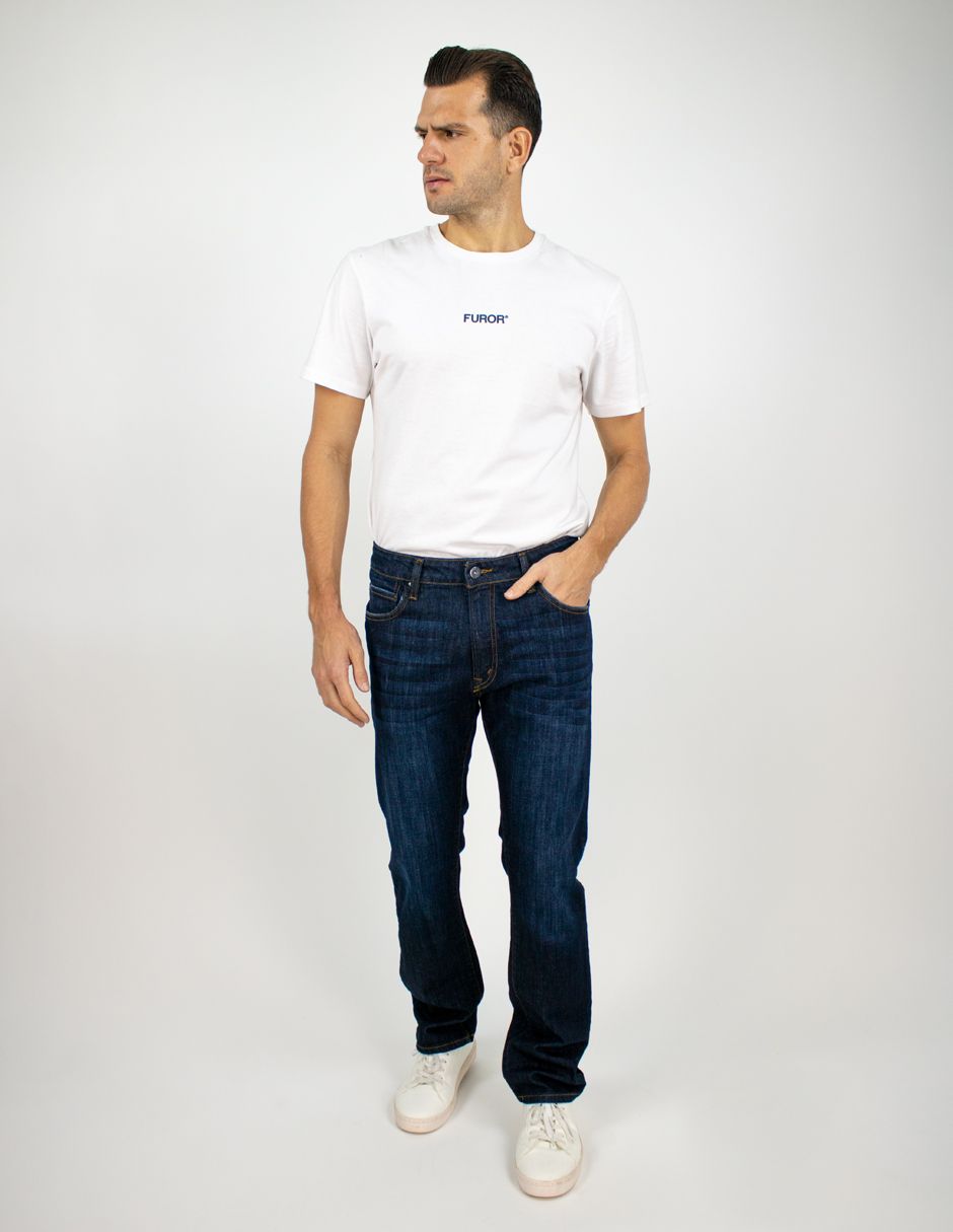 Hombre con camiseta blanca y vaqueros oscuros de pie sobre un fondo claro, mirando hacia un lado.