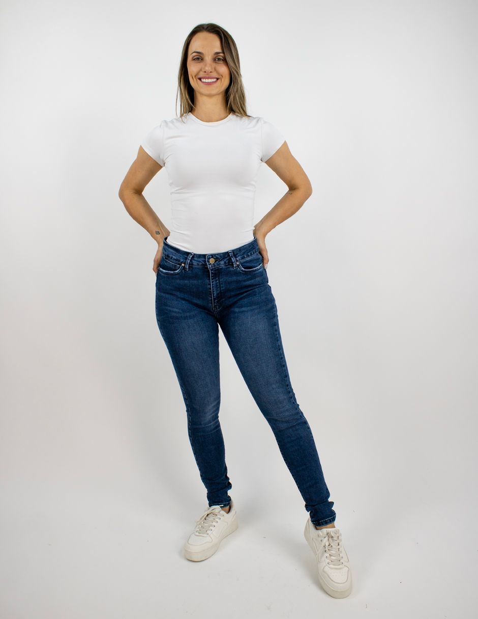 Mujer de pie sobre un fondo blanco, vestida con una camiseta blanca, vaqueros azules y zapatillas blancas.