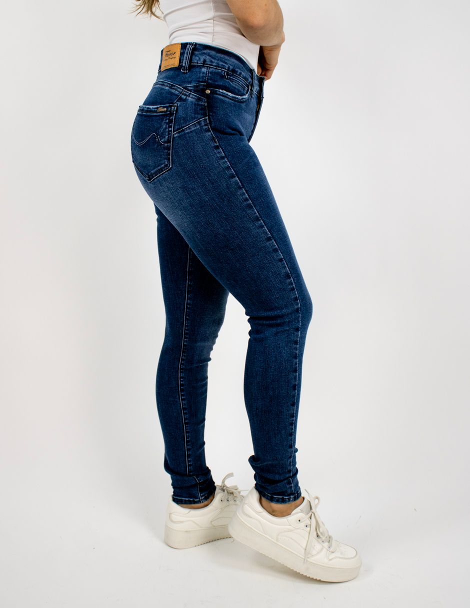 Vista lateral de una persona con vaqueros azul oscuro, camiseta beige y zapatillas blancas sobre un fondo blanco.