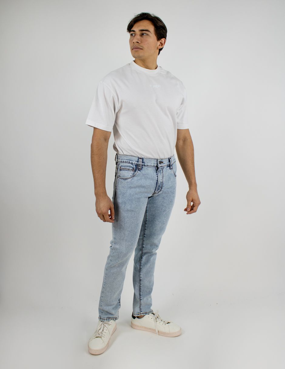 Persona con camiseta blanca, vaqueros azul claro y zapatillas blancas de pie sobre un fondo blanco liso.