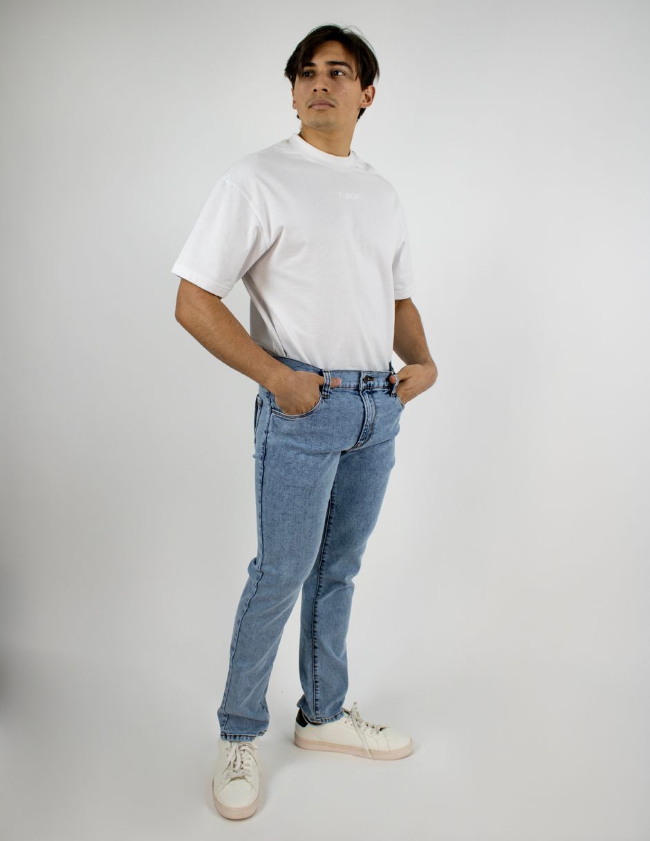 De pie, con una camiseta blanca, vaqueros azul claro y zapatillas blancas, sobre un fondo blanco liso.