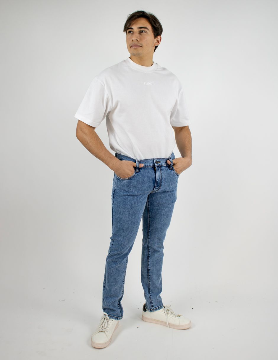 Hombre de pie con camiseta blanca, vaqueros azules y zapatillas blancas sobre un fondo blanco liso.