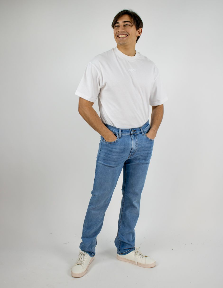 De pie, sonriendo, con una camiseta blanca, vaqueros azules y zapatillas blancas sobre un fondo gris liso.