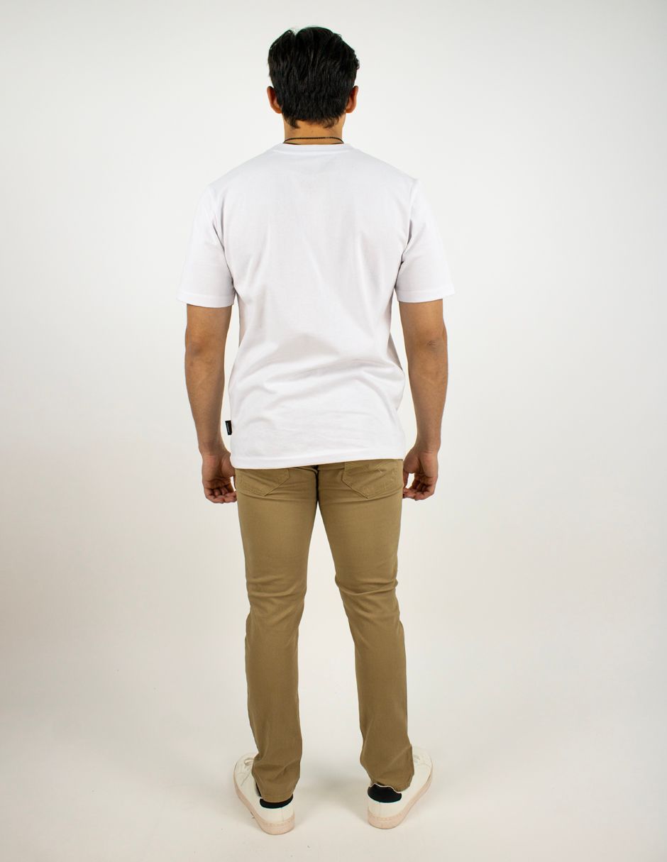 Persona de pie de espaldas a la cámara, con camiseta blanca, pantalones color canela y zapatos blancos, sobre un fondo liso.