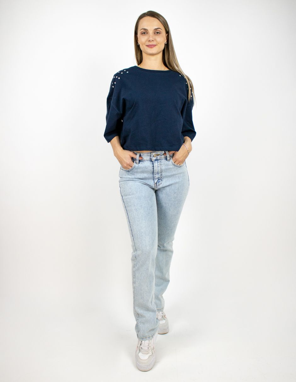 Mujer de pie con suéter azul marino, jeans claros y zapatillas blancas sobre un fondo blanco.