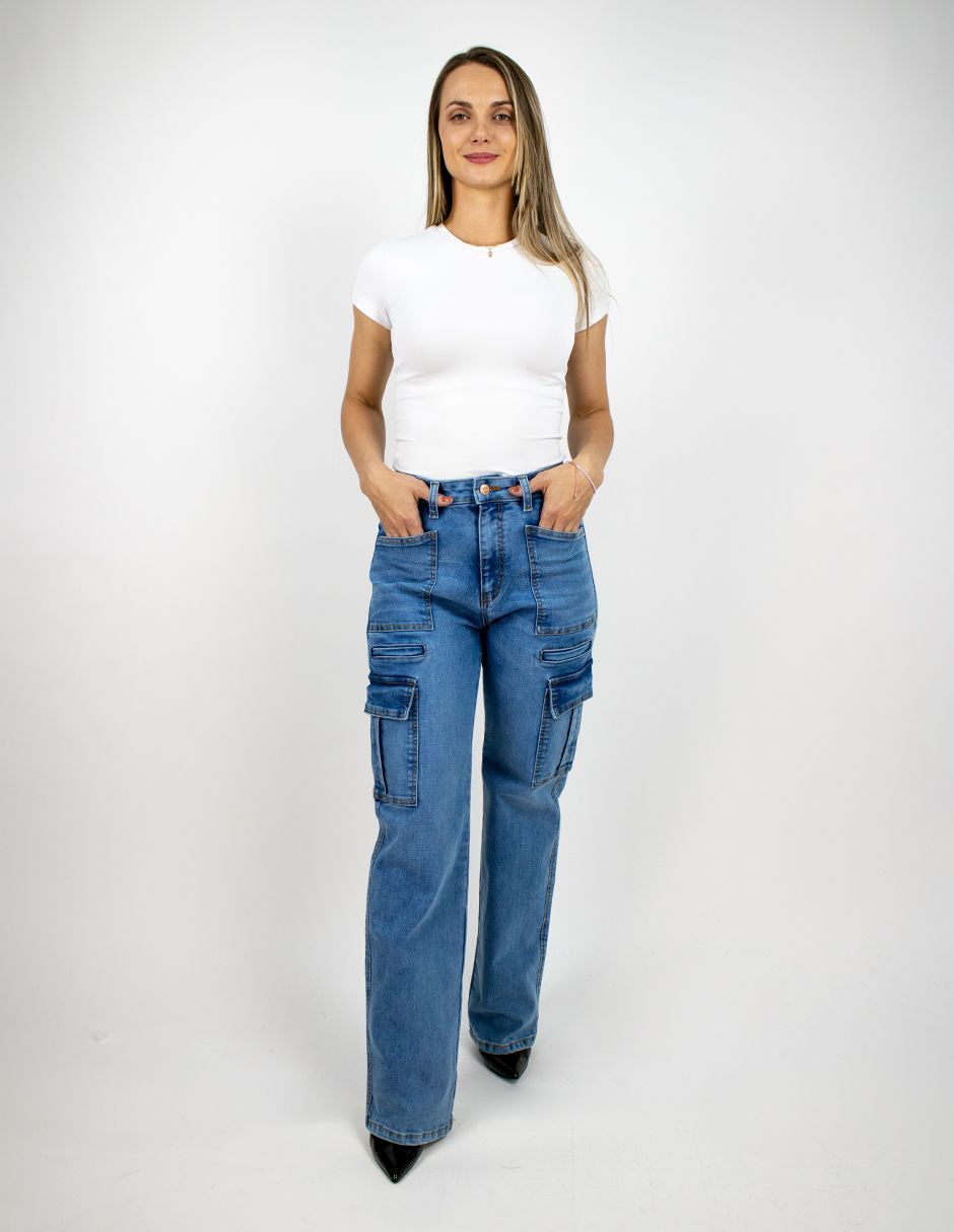 Mujer con blusa blanca y pantalones vaqueros cargo azules, de pie frente a un fondo claro liso.