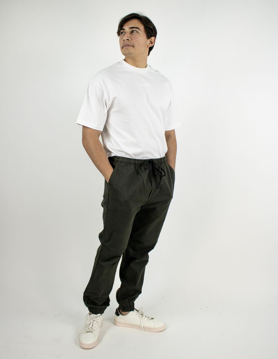 Persona de pie con una camiseta blanca, pantalones oscuros y zapatillas blancas sobre un fondo de estudio liso.