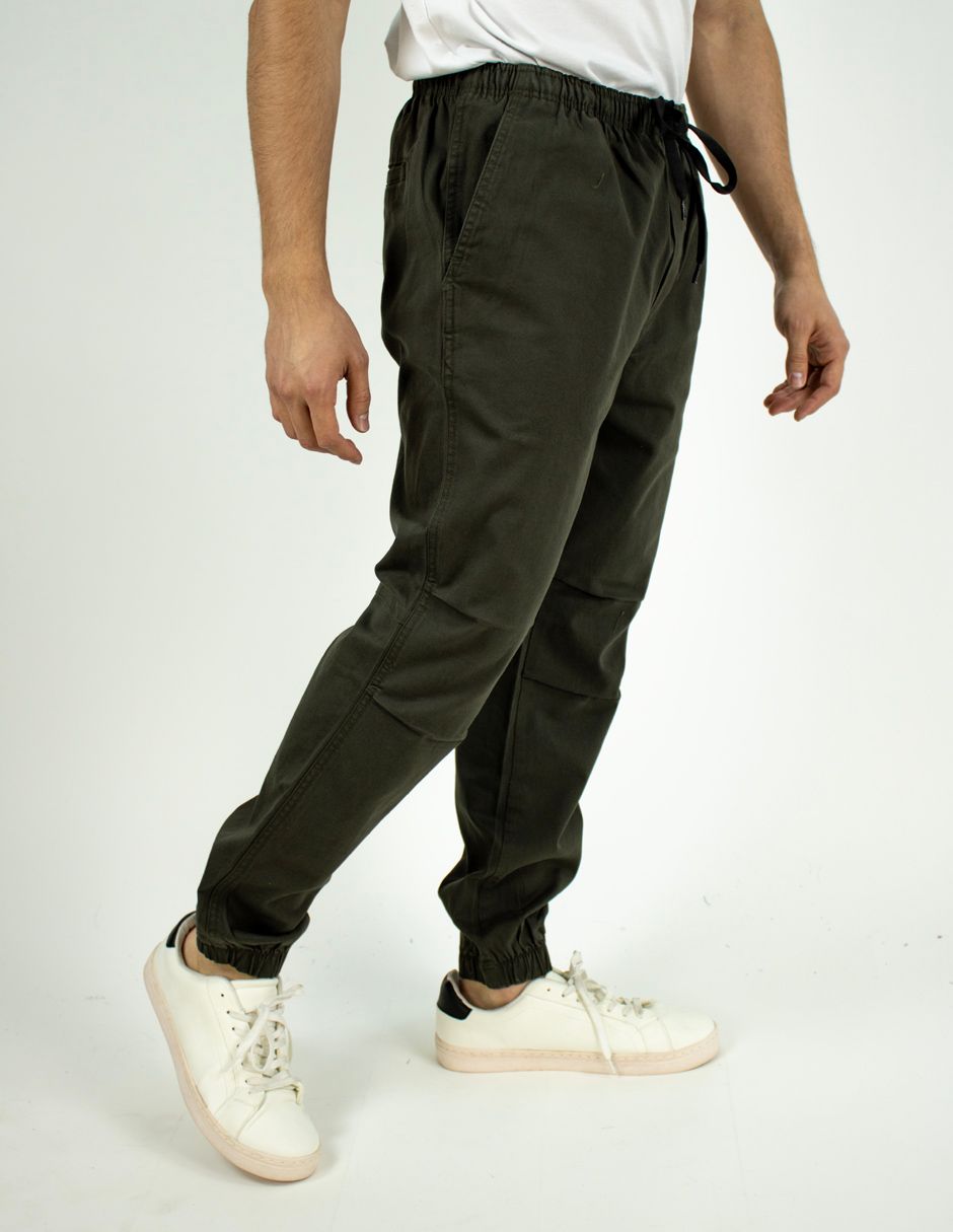Persona con pantalones deportivos verde oscuro y zapatillas blancas, vista de perfil sobre un fondo liso.