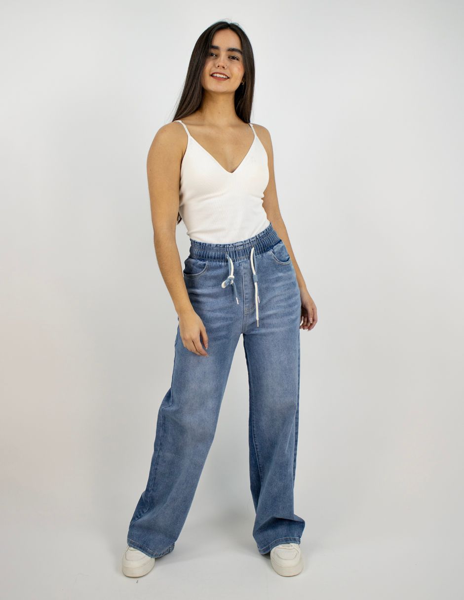 Mujer de pie con una camiseta blanca sin mangas, vaqueros azules de pierna ancha y zapatillas blancas sobre un fondo liso.