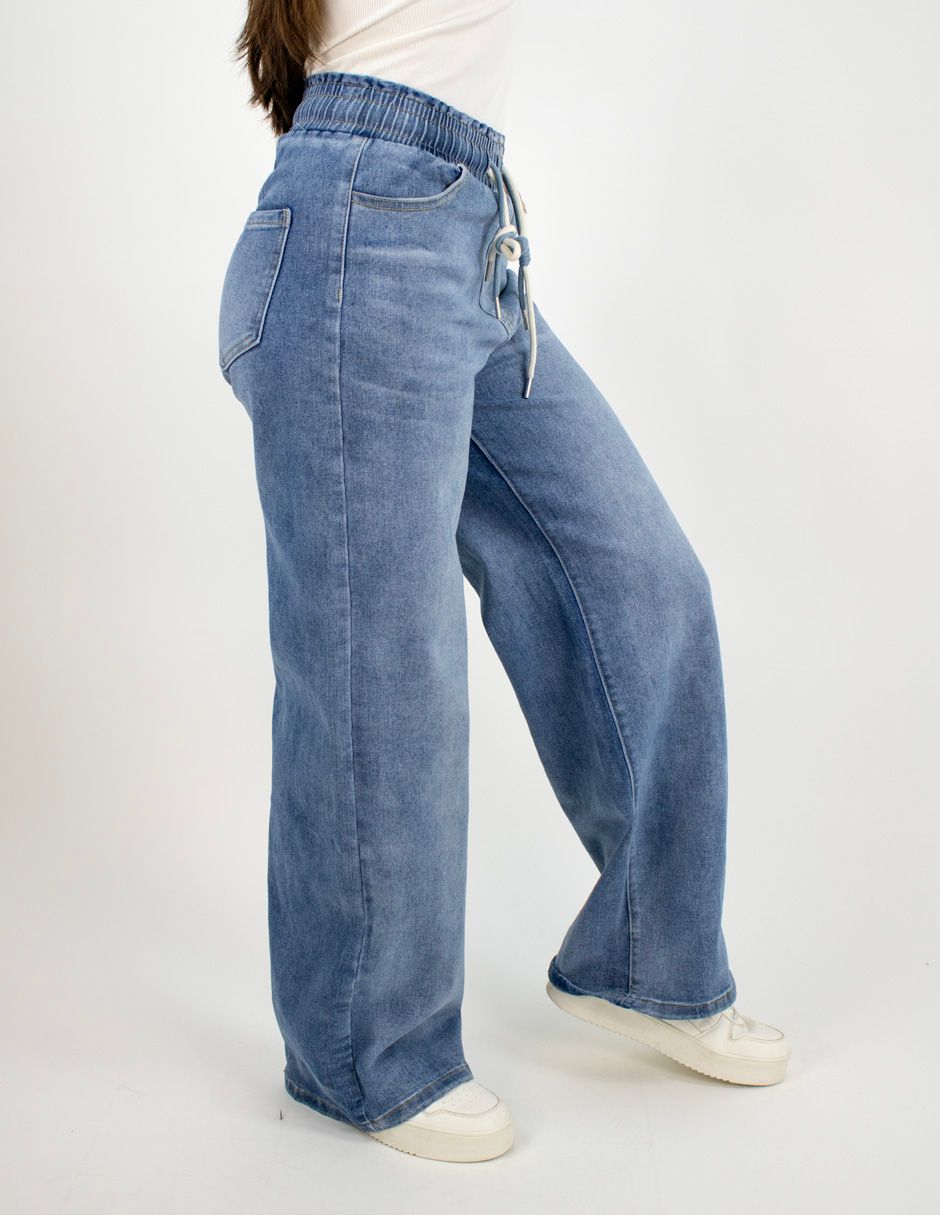 Persona con pantalones vaqueros azules de pierna ancha y zapatillas blancas sobre un fondo blanco liso.