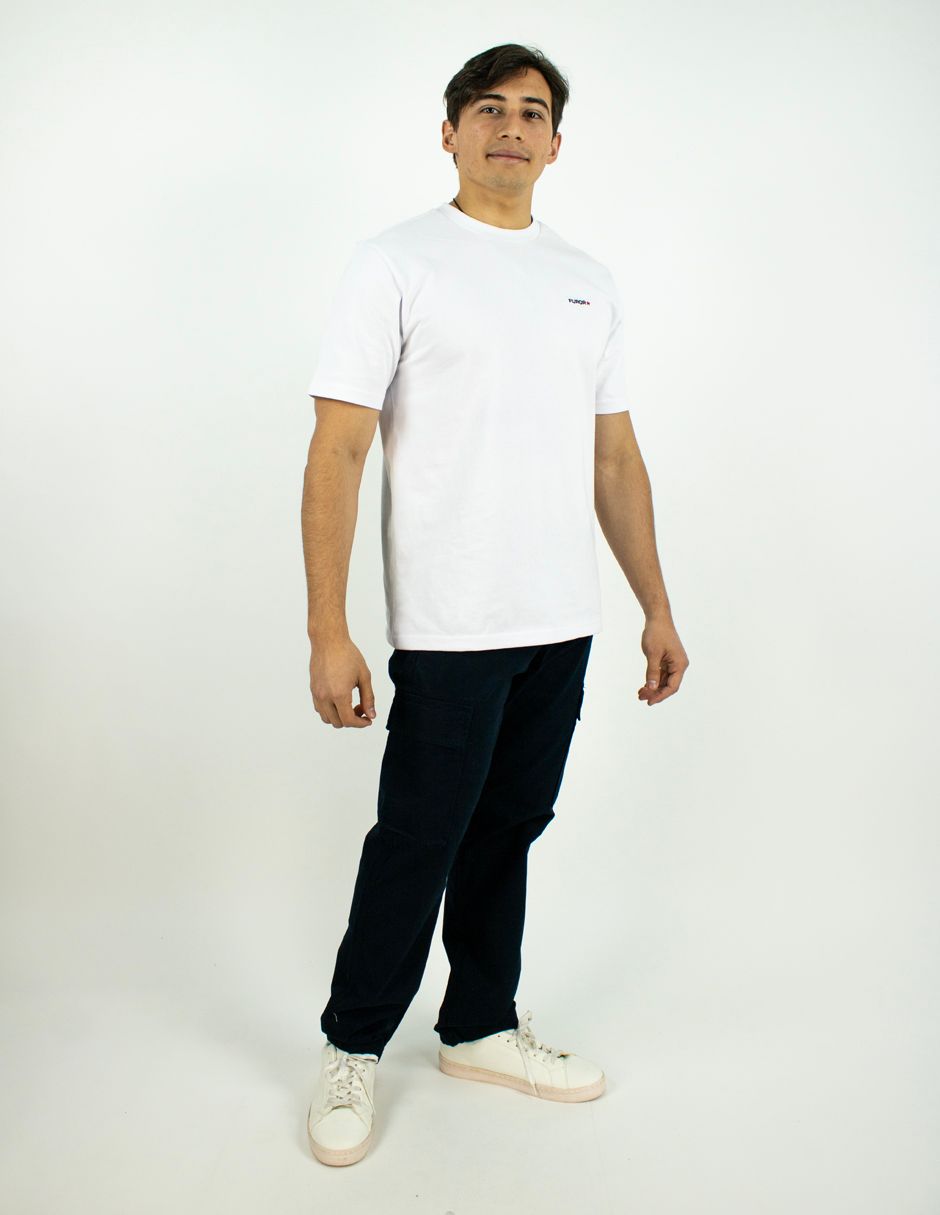 Persona de pie sobre un fondo blanco, vestida con una camiseta blanca, pantalones oscuros y zapatillas blancas.