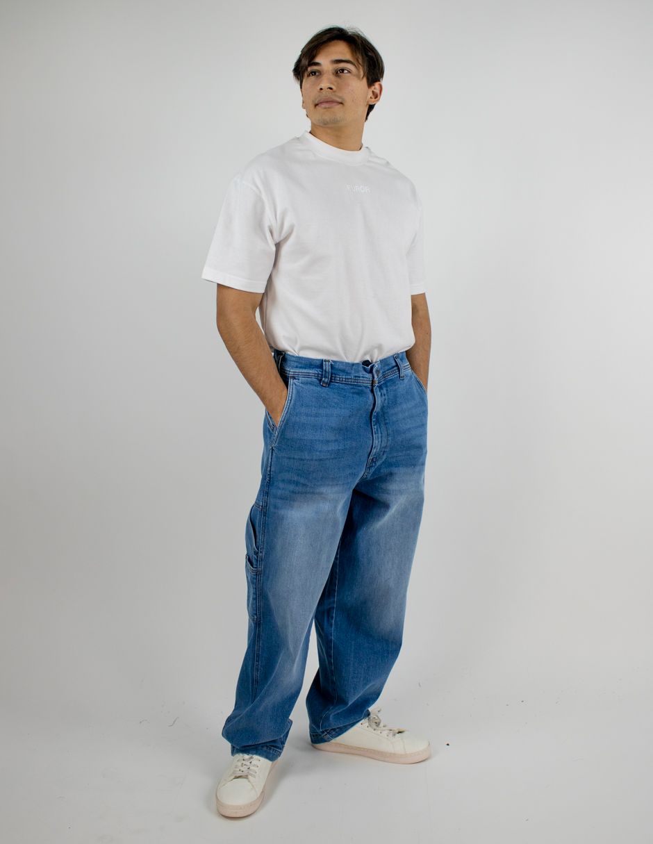 Persona de pie con camiseta blanca, vaqueros azules y zapatillas blancas sobre un fondo claro liso.