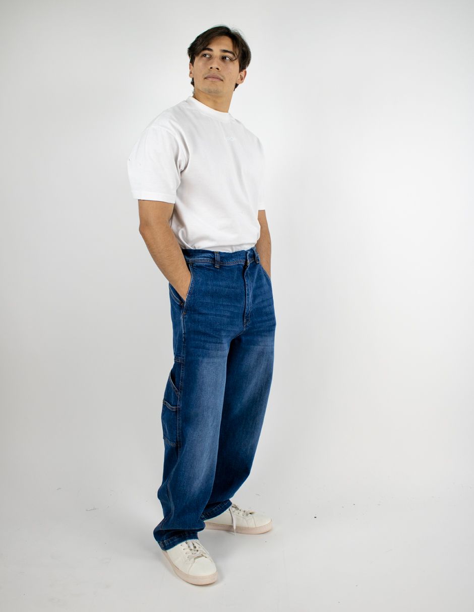 Hombre de pie frente a un fondo blanco, vistiendo una camisa blanca, vaqueros azules y zapatillas blancas.