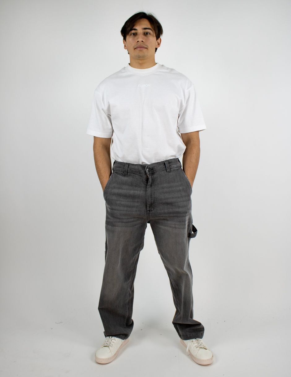 Persona de pie sobre un fondo blanco, vestida con una camiseta blanca, vaqueros grises y zapatillas blancas.