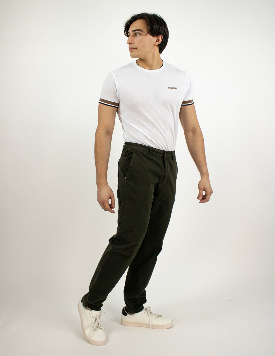 Hombre de pie con camiseta blanca, pantalones oscuros y zapatillas blancas sobre un fondo liso.