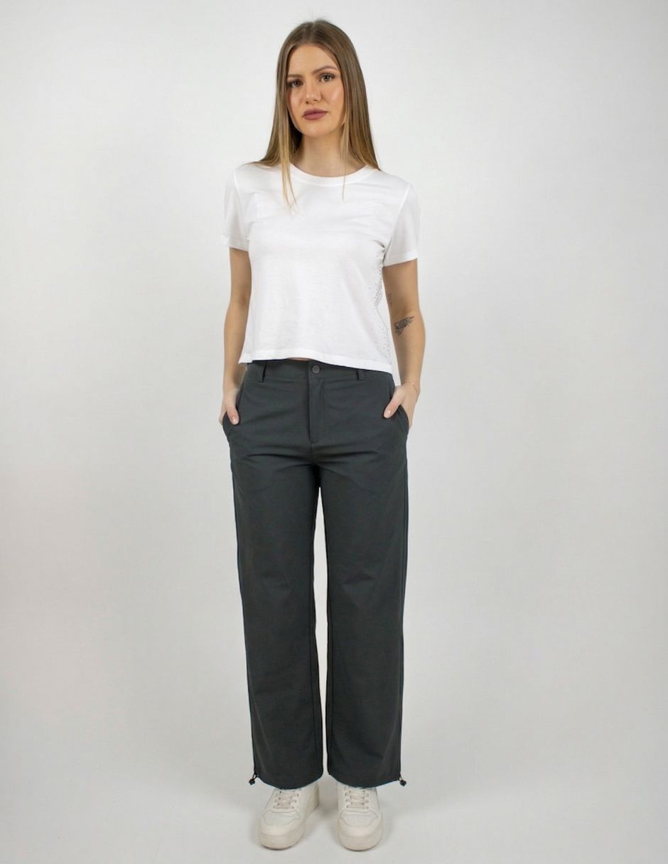 Mujer de pie con camiseta blanca y pantalones oscuros sobre un fondo liso.