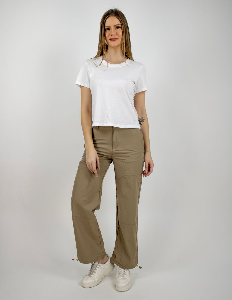 Mujer de pie con camiseta blanca, pantalones verde oliva y zapatillas blancas sobre un fondo gris liso.