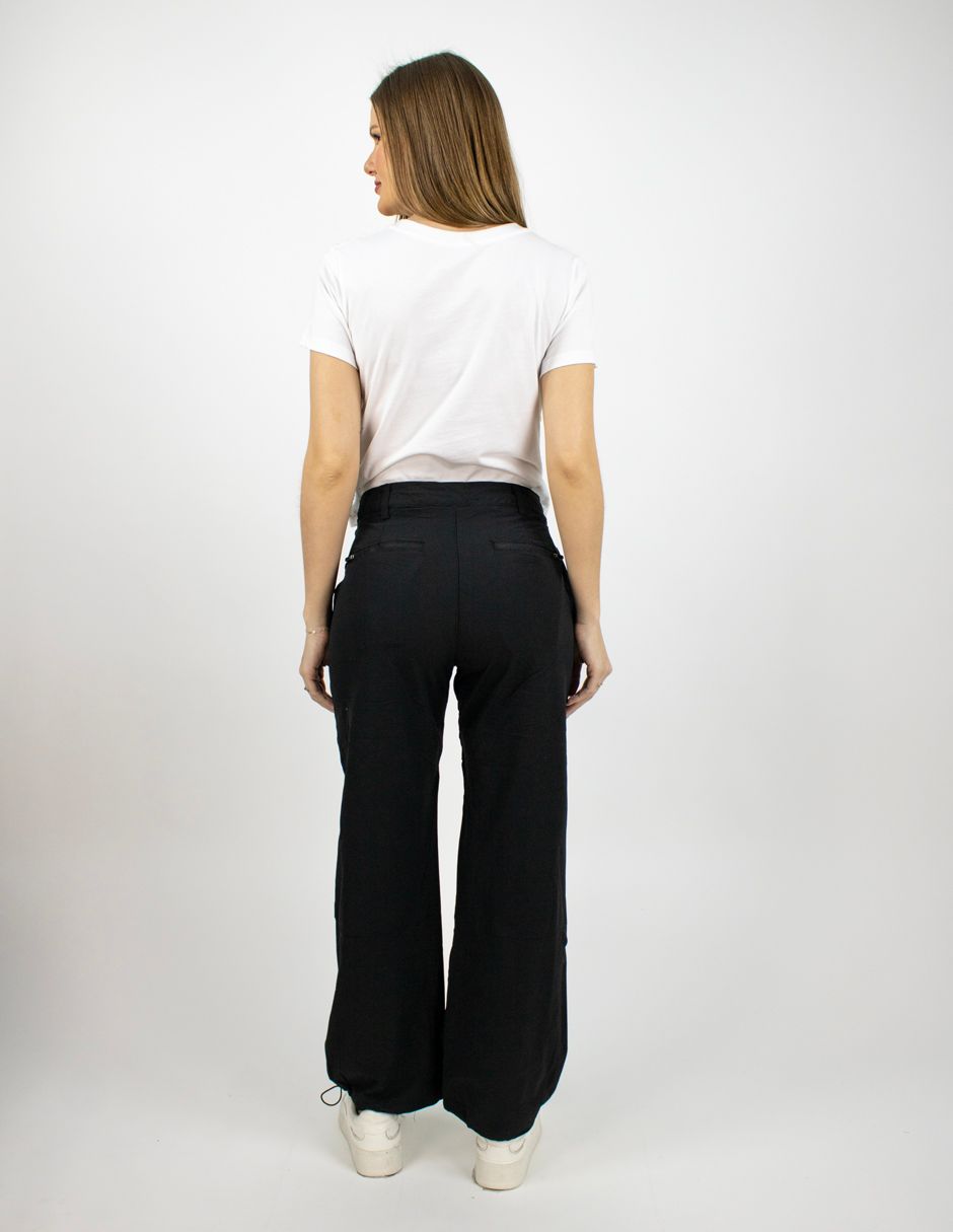 Persona de pie de espaldas a la cámara, con camiseta blanca, pantalones negros y zapatos blancos, sobre un fondo liso.