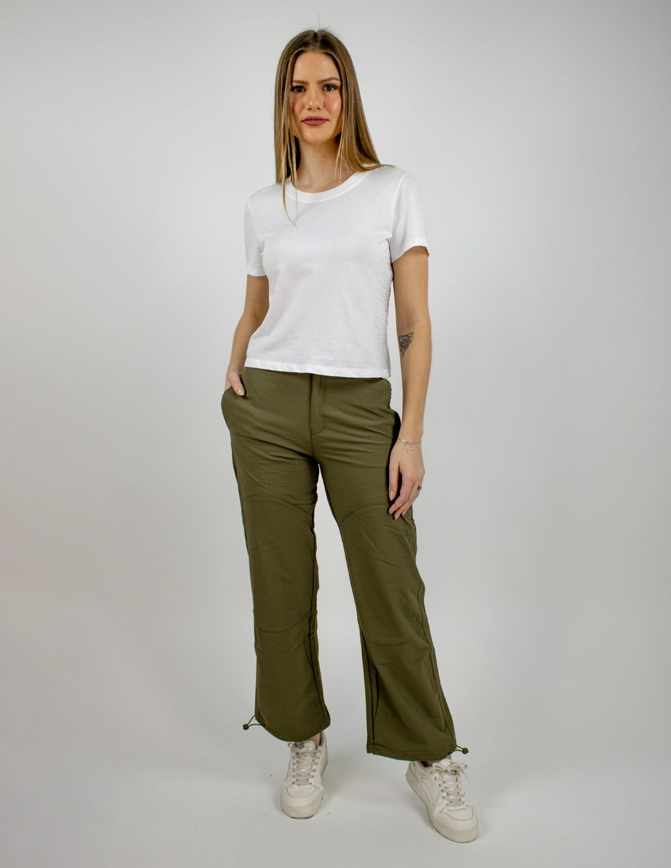 Mujer de pie con una camiseta blanca, pantalones verde oliva y zapatillas blancas sobre un fondo liso.