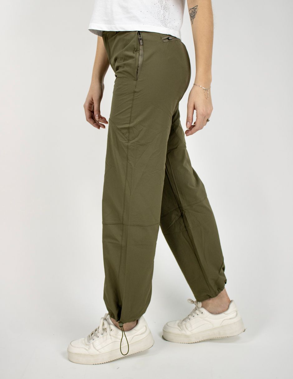Persona con pantalones verde oliva y zapatillas blancas, de pie de perfil sobre un fondo liso.