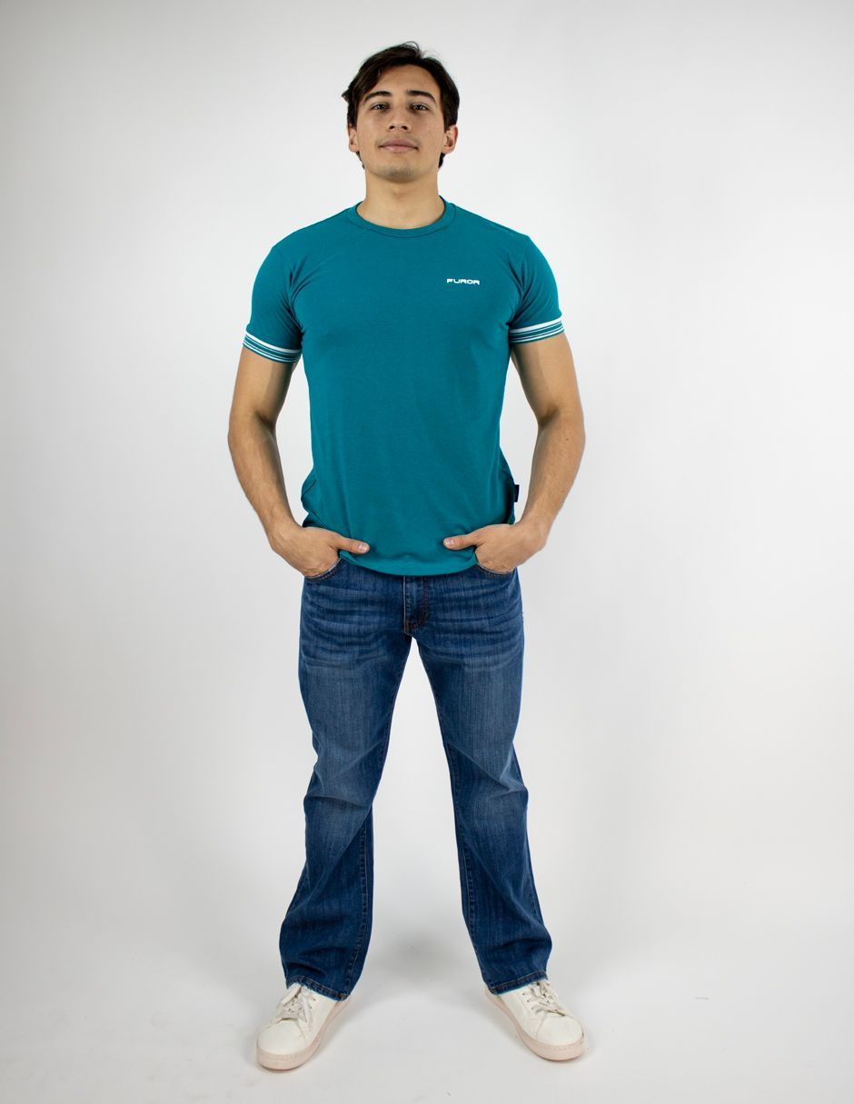 Hombre de pie sobre un fondo blanco, con camiseta color turquesa, vaqueros y zapatillas blancas.
