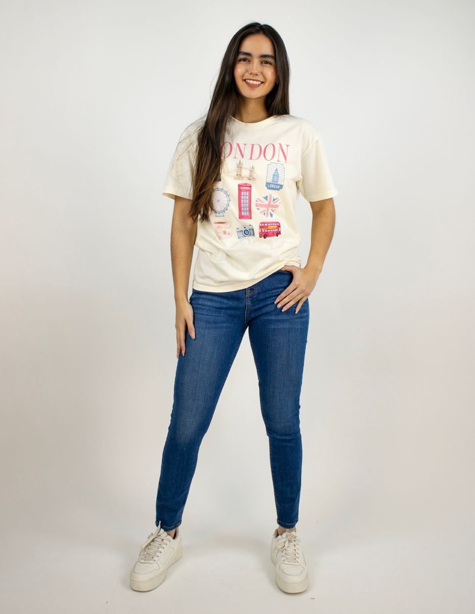 Mujer de pie sobre un fondo blanco, vestida con una camiseta estampada, vaqueros azules y zapatillas blancas.