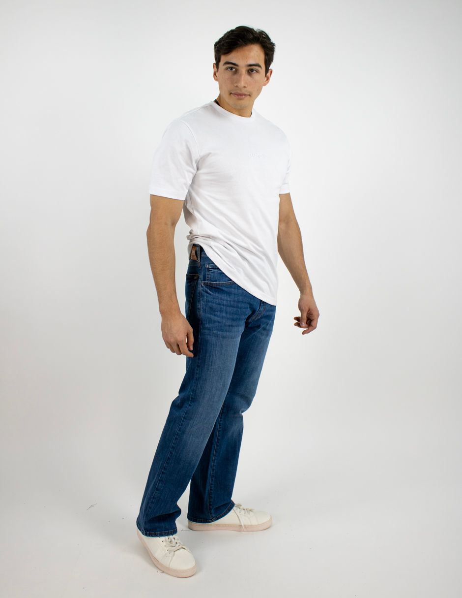 Hombre de pie con camiseta blanca, vaqueros azules y zapatillas blancas sobre un fondo blanco liso.