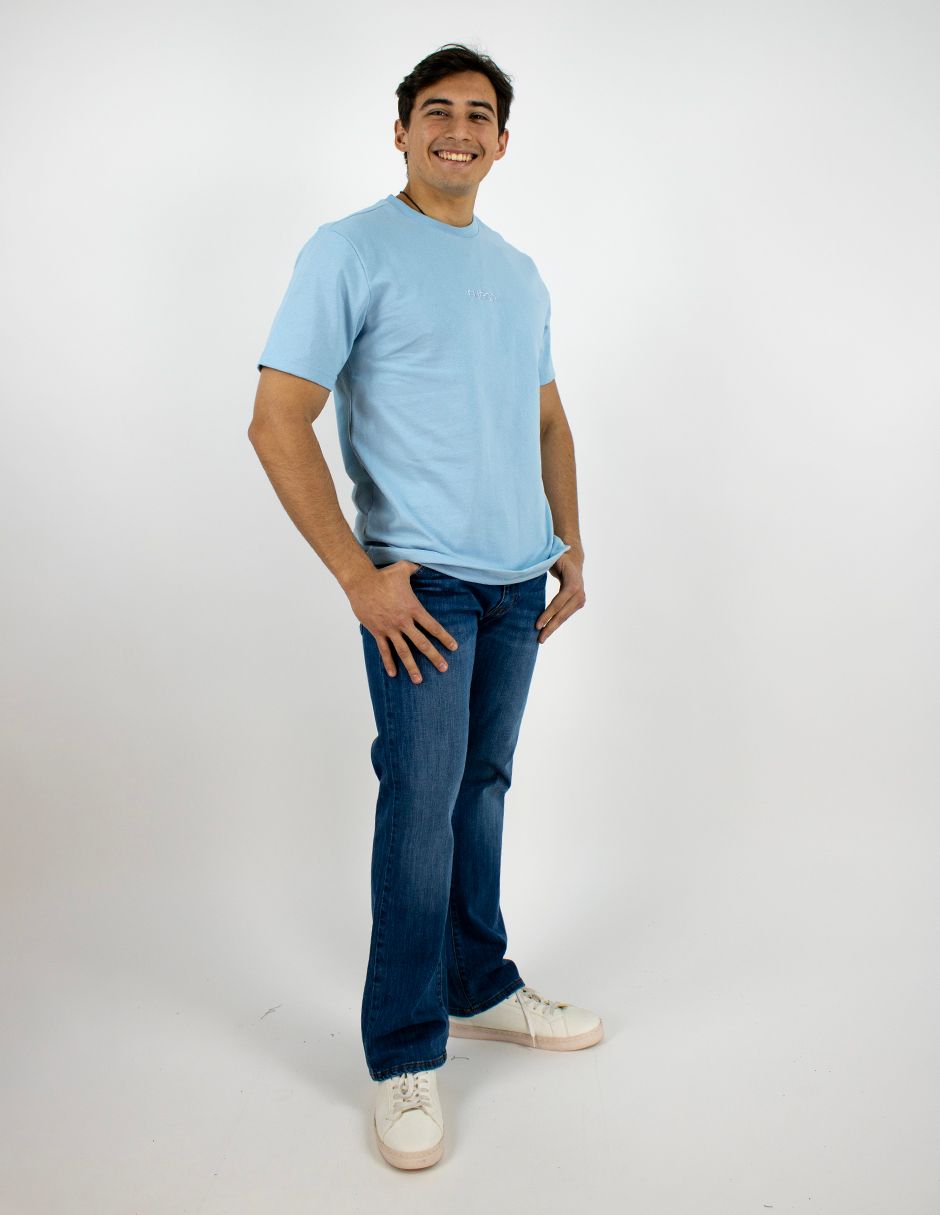 Hombre con camiseta azul claro y vaqueros, de pie sobre un fondo blanco, sonriendo.