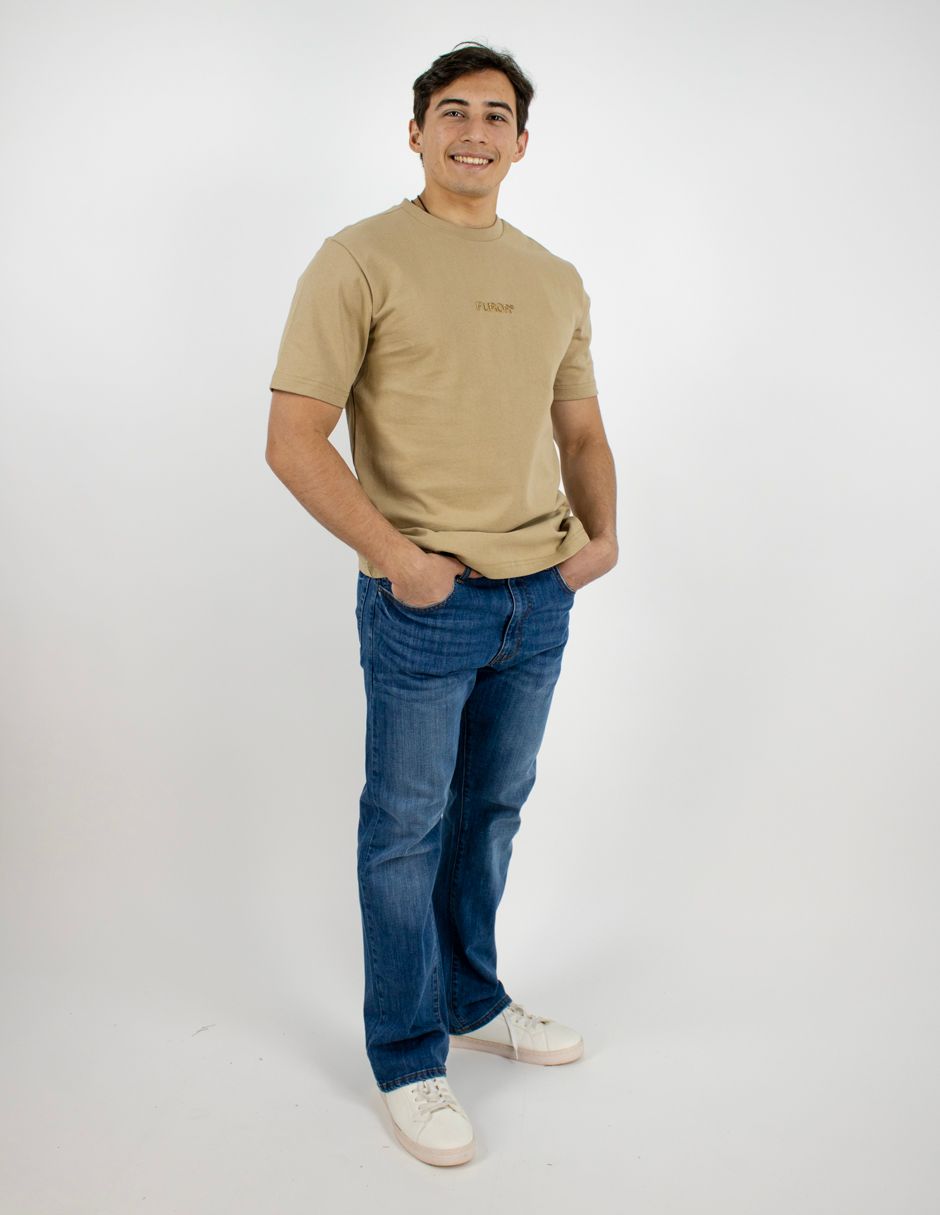 De pie, con un aire informal, frente a un fondo blanco, viste una camiseta color canela, vaqueros azules y zapatillas blancas.