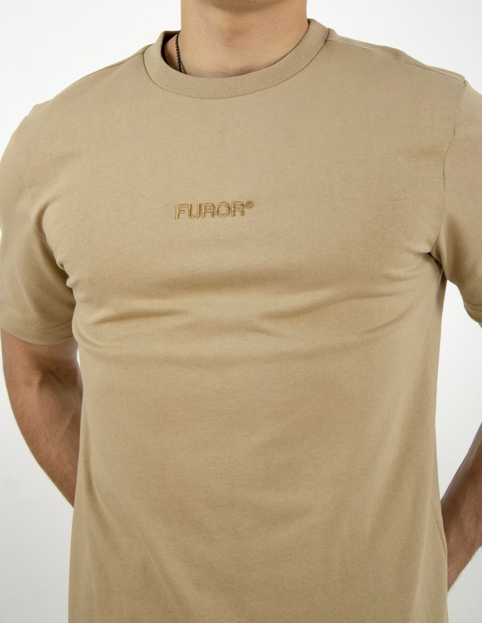 Camiseta beige con un pequeño logo en el pecho, mostrada en una persona sobre un fondo liso.