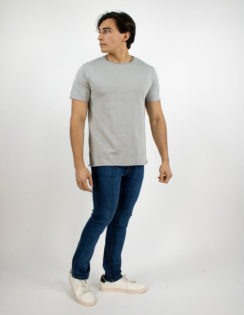 Hombre con camiseta gris, vaqueros azules y zapatillas blancas de pie sobre un fondo blanco liso.