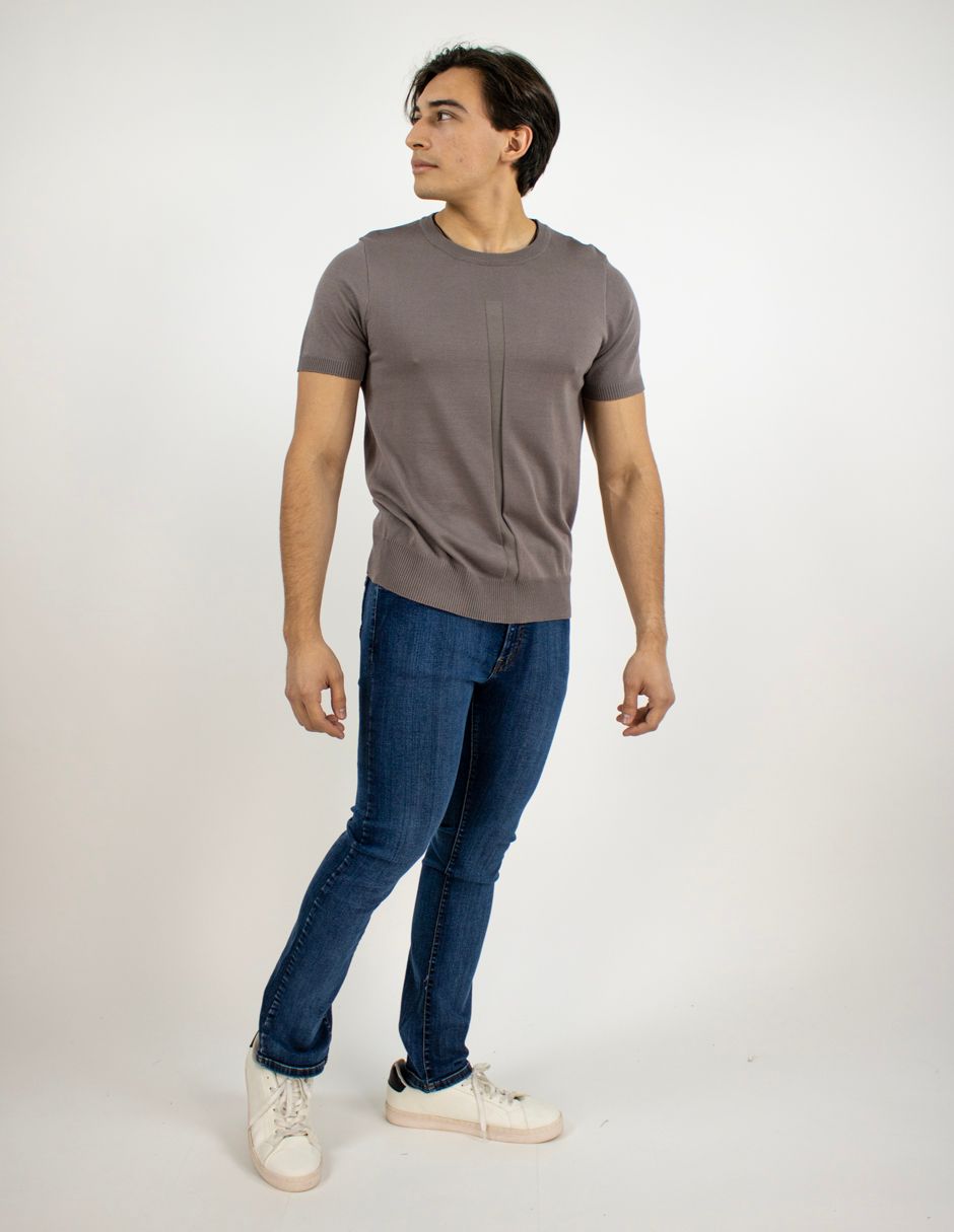 Hombre de pie con una camiseta gris, vaqueros azules y zapatillas blancas sobre un fondo blanco liso.
