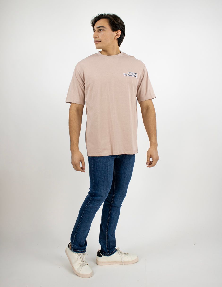 Hombre de pie con una camiseta beige, vaqueros azules y zapatillas blancas sobre un fondo blanco liso.
