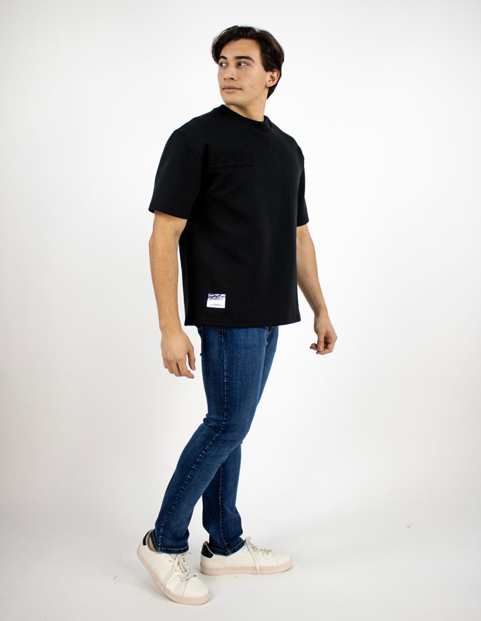 Persona de pie con una camiseta negra, vaqueros azules y zapatillas blancas sobre un fondo blanco.