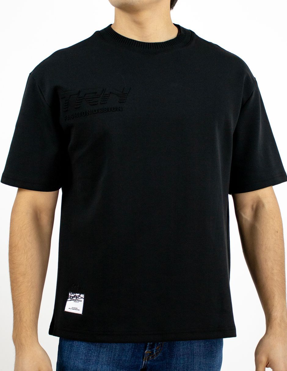 Camiseta negra puesta a una persona, vista frontal, con un pequeño parche con el logo cerca del dobladillo.