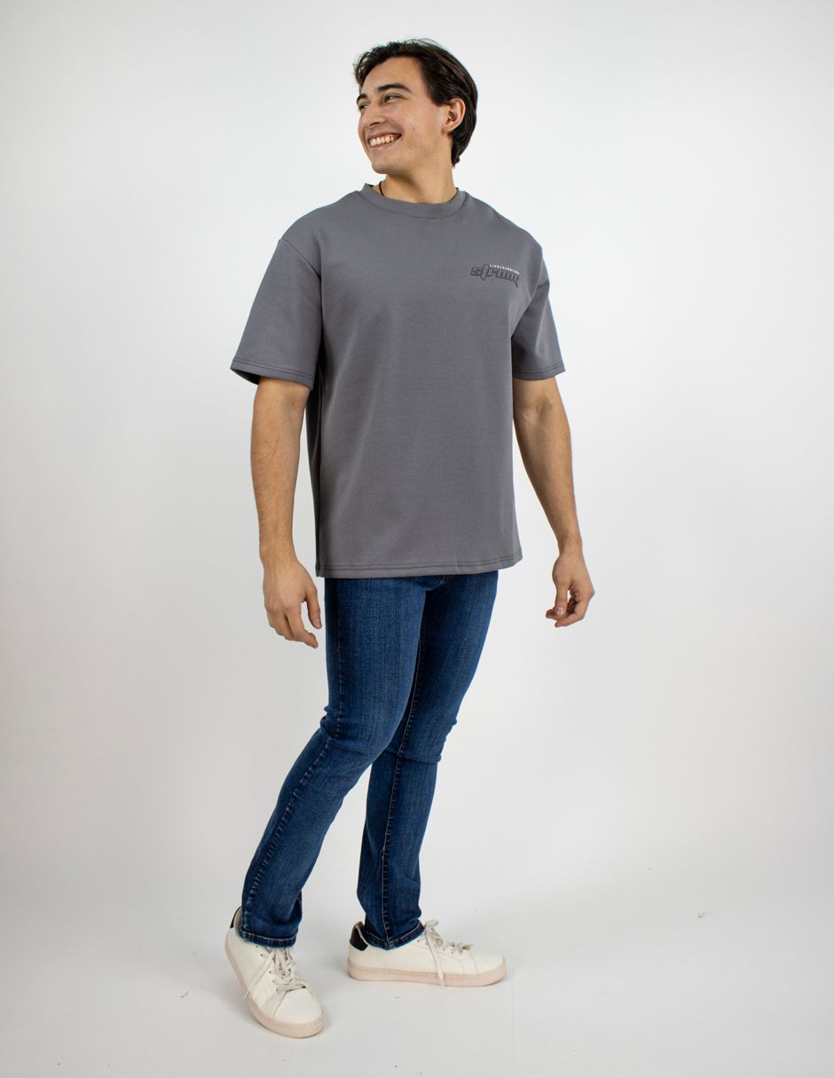 Persona de pie sobre un fondo blanco, vestida con una camiseta gris, vaqueros azules y zapatillas blancas, mirando hacia la izquierda y sonriendo.