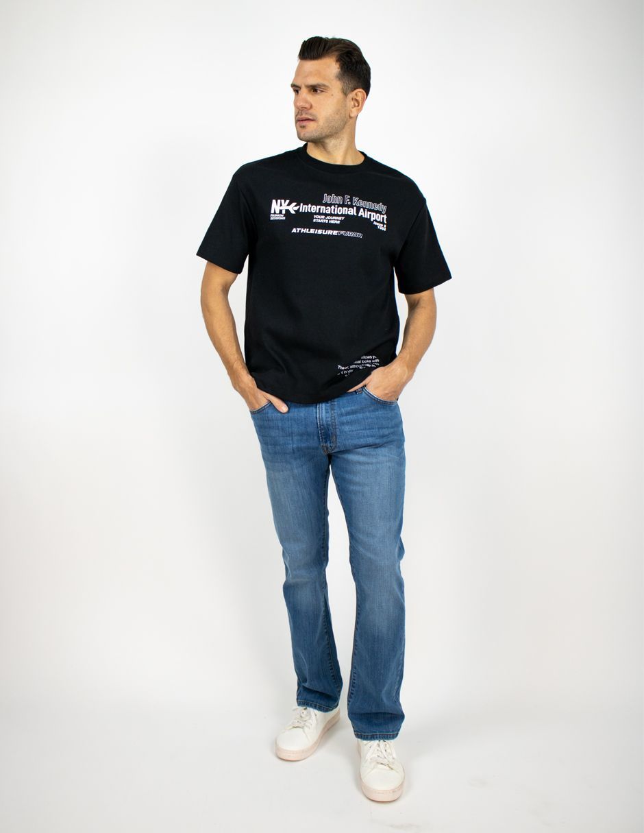 Hombre de pie con camiseta negra y vaqueros, con las manos en los bolsillos, mirando hacia un lado sobre un fondo blanco.