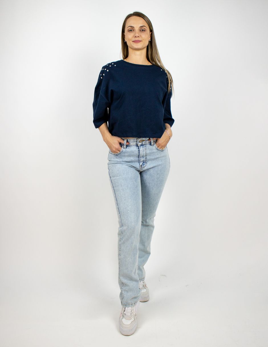 Mujer de pie con una blusa azul marino, vaqueros claros y zapatillas blancas sobre un fondo blanco.