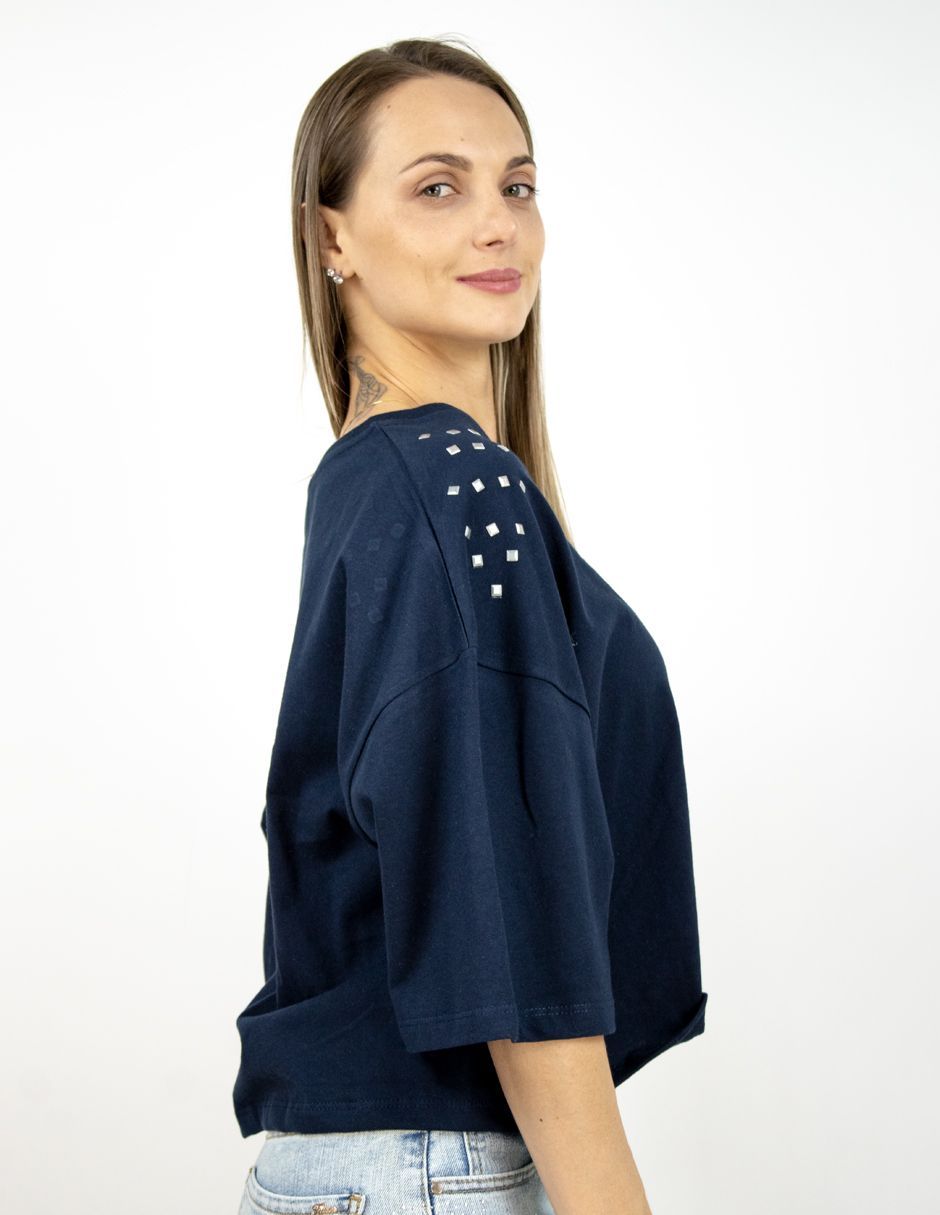 Mujer con top azul marino con hombreras tachonadas, mostrada de perfil lateral sobre un fondo blanco.