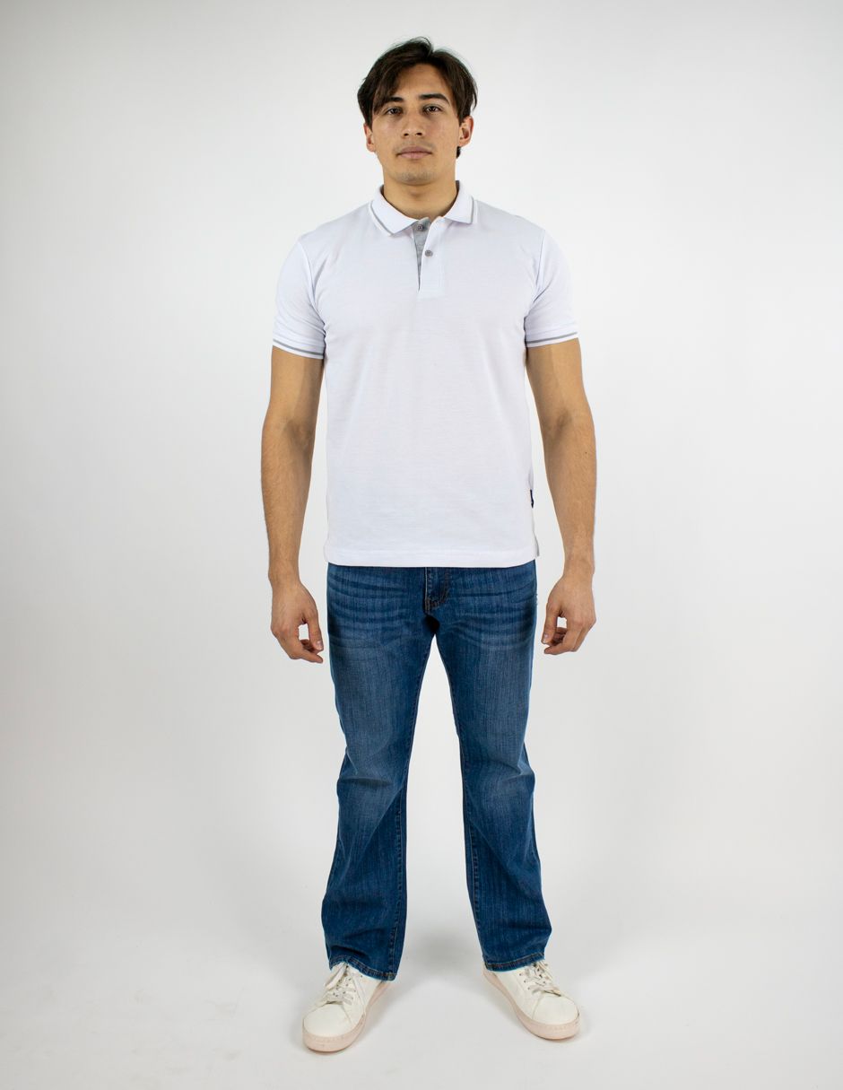 Hombre de pie sobre un fondo blanco, vistiendo una camisa polo blanca, vaqueros azules y zapatillas blancas.